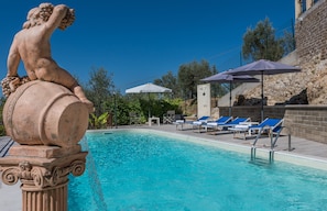 Une piscine ornée d’une statue représentant une figure tenant une grappe de raisins, entourée de chaises longues et de parasols.