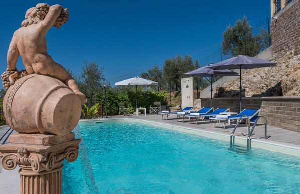 Une piscine ornée d’une statue représentant une figure tenant une grappe de raisins, entourée de chaises longues et de parasols.