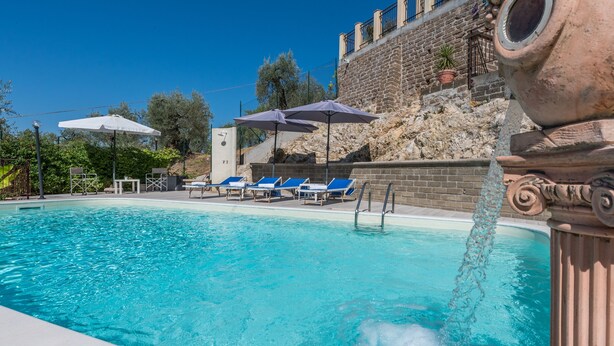 Une piscine d’un bleu éclatant, bordée de chaises longues et d’ombrelles, et entourée de murs de pierre et de verdure.