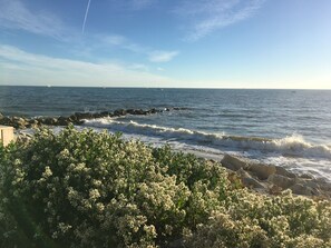 A coastal view with waves crashing onto rocks, a clear sky, and distant sailboats.