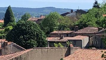A view of a town with terracotta-roofed buildings, surrounded by greenery and hills in the background.