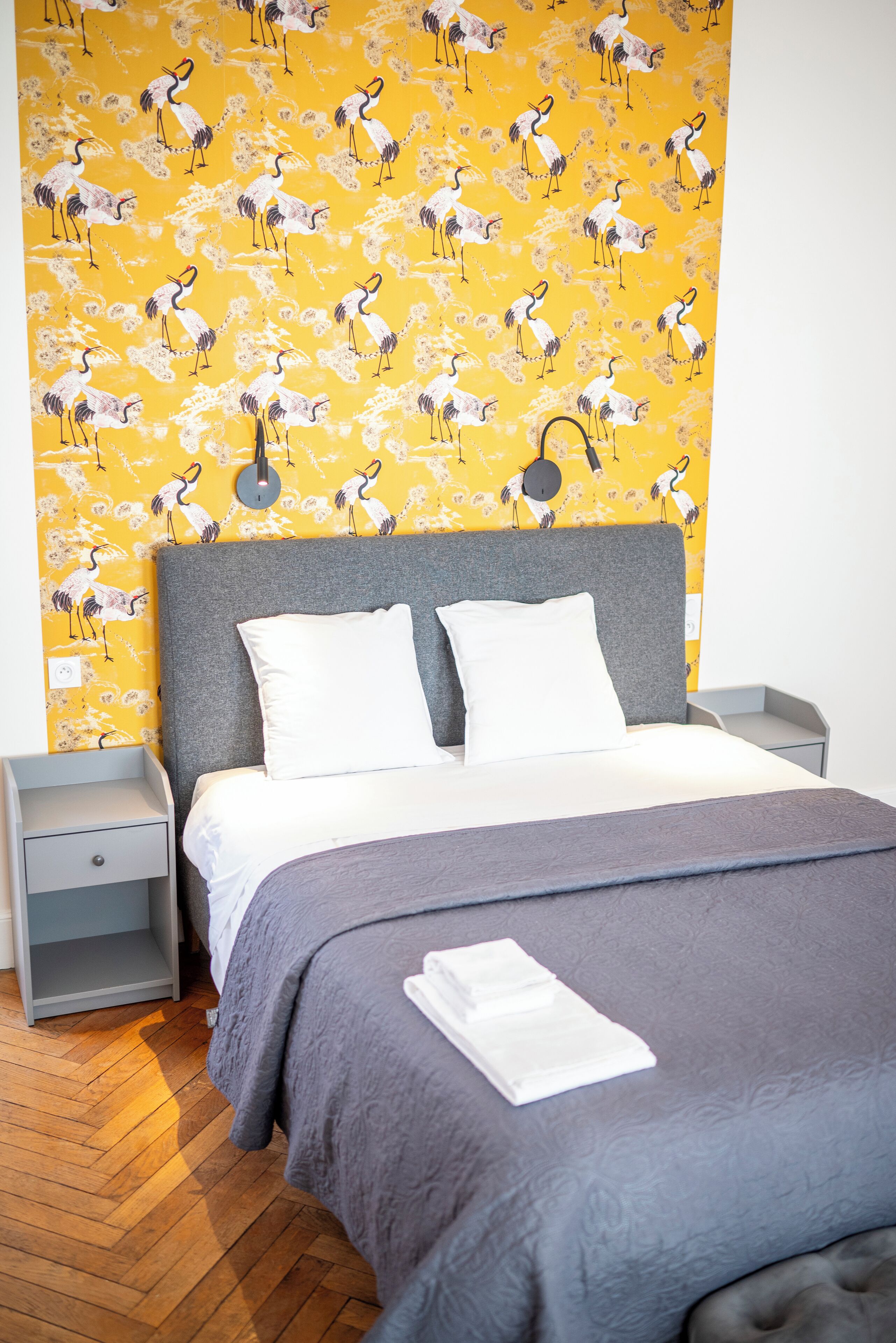 A neatly made bed with a grey headboard and white pillows, a grey nightstand, and a wall with a yellow wallpaper featuring a pattern of birds.