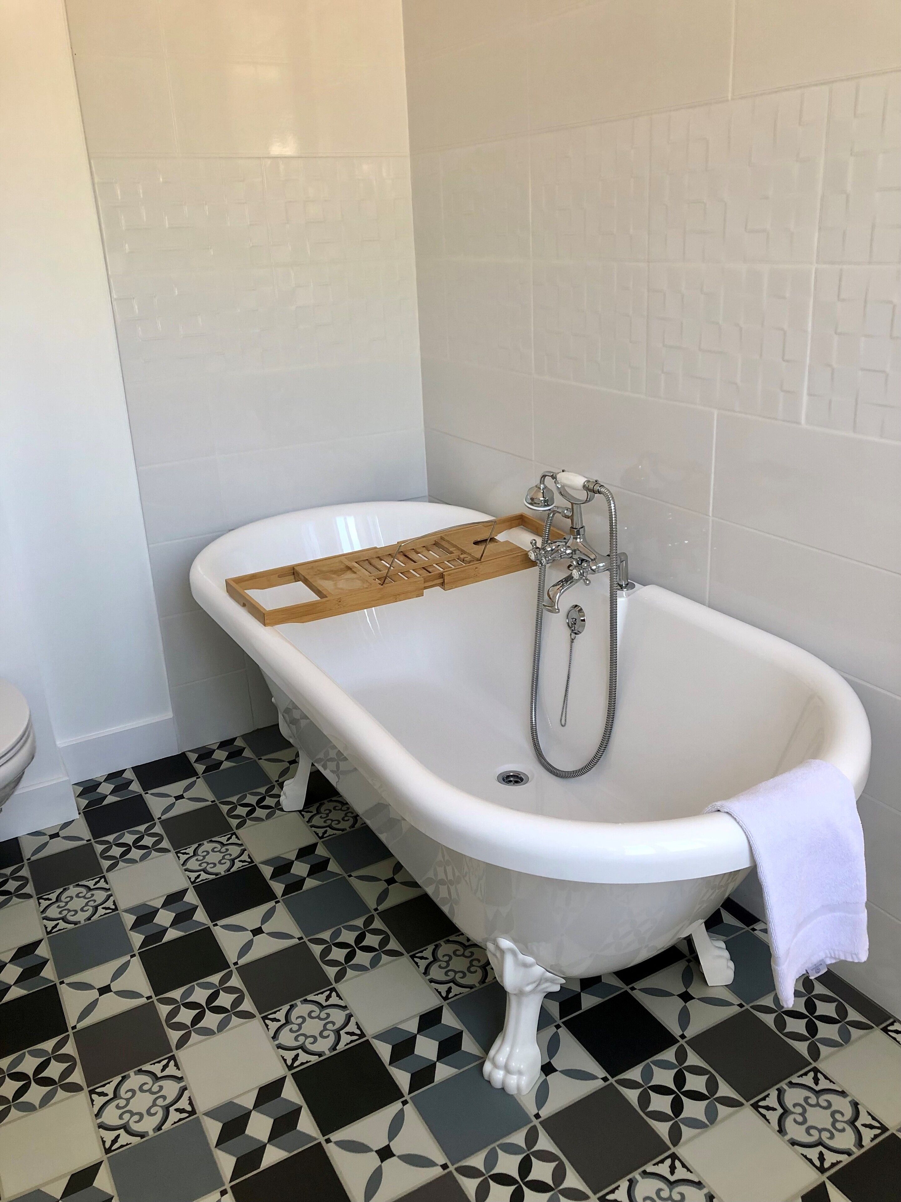 A bathroom with a white bathtub, a wooden tray, a showerhead, and patterned floor tiles.