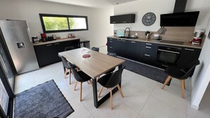 A modern kitchen with a dining area, featuring a refrigerator, stove, sink, and a large window. - Single-storey house Royan 8 pers - Heated swimming pool in summer - Pontaillac beach (VAUX SUR MER)