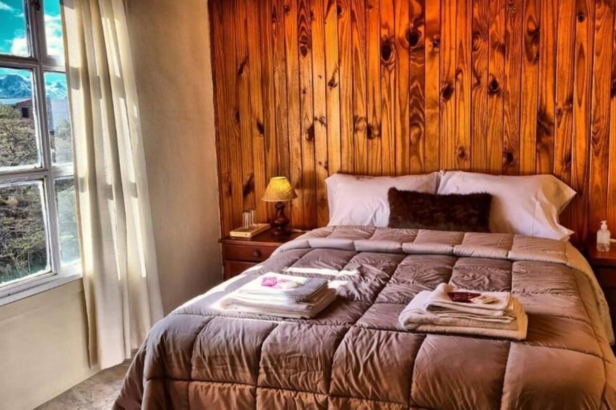 A bedroom with a wooden headboard, a bed with a brown quilt, two bedside tables with lamps, and a window with curtains.