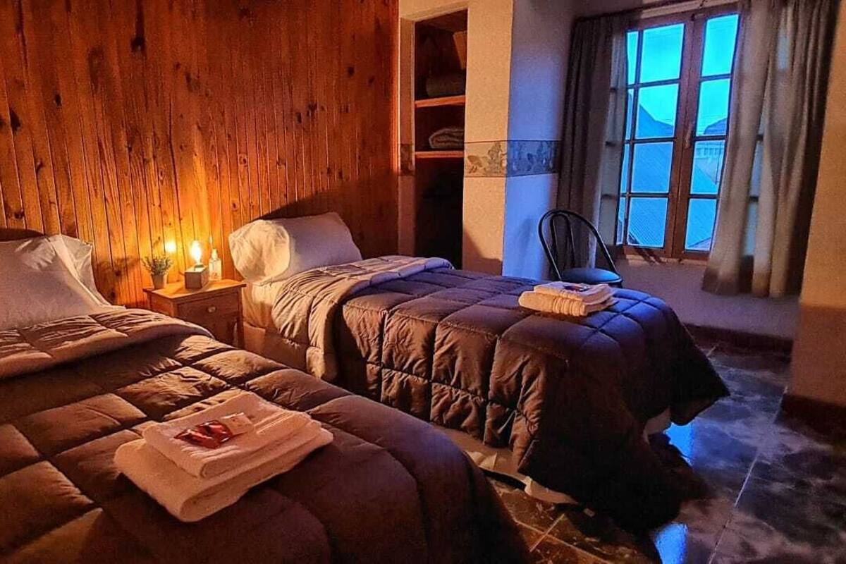 A cozy room with two beds, wooden walls, a window with curtains, and a nightstand with a lamp.
