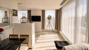 A modern living room with a dining area, a flat-screen TV, a tripod, and large windows with curtains.