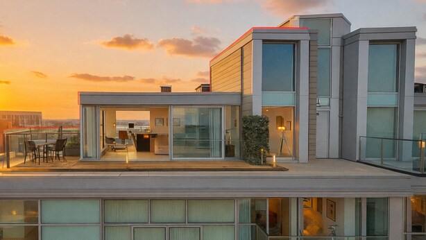 Un edificio moderno con fachada de vidrio, una terraza en la azotea con mobiliario exterior y una vista despejada del atardecer.