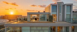 A modern building with a glass facade, a rooftop terrace with outdoor furniture, and a clear view of the sunset.