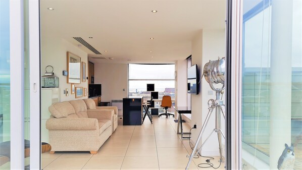 A modern living room with a sofa, a desk, and a television. There is a large window with a view of the city.