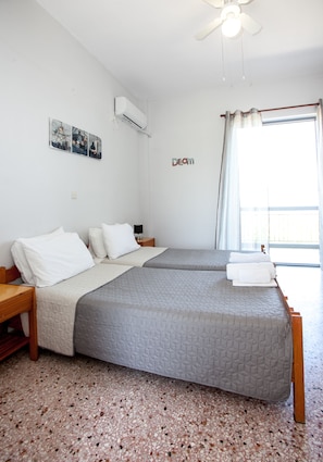 A hotel room with two beds, a ceiling fan, an air conditioning unit, a window with curtains, and a small table with a lamp.