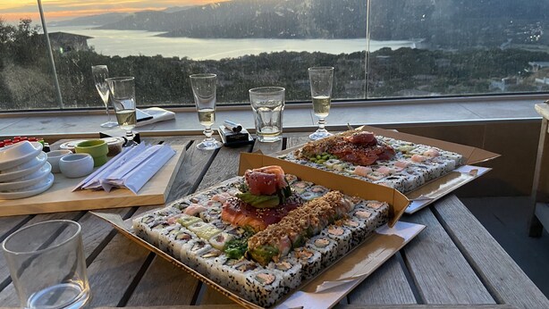 A table with sushi boxes and glasses of white wine, overlooking a scenic view of mountains and a lake. - Villa Propriano 6 to 8 people VILLA WITH POOL AND SEA VIEW (VIGGIANELLO)