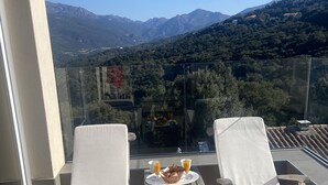 A balcony with two white cushioned chairs, a small table with a basket of croissants and two glasses of orange juice, and a scenic view of mountains and greenery. - Villa Propriano 6 to 8 people VILLA WITH POOL AND SEA VIEW (VIGGIANELLO)