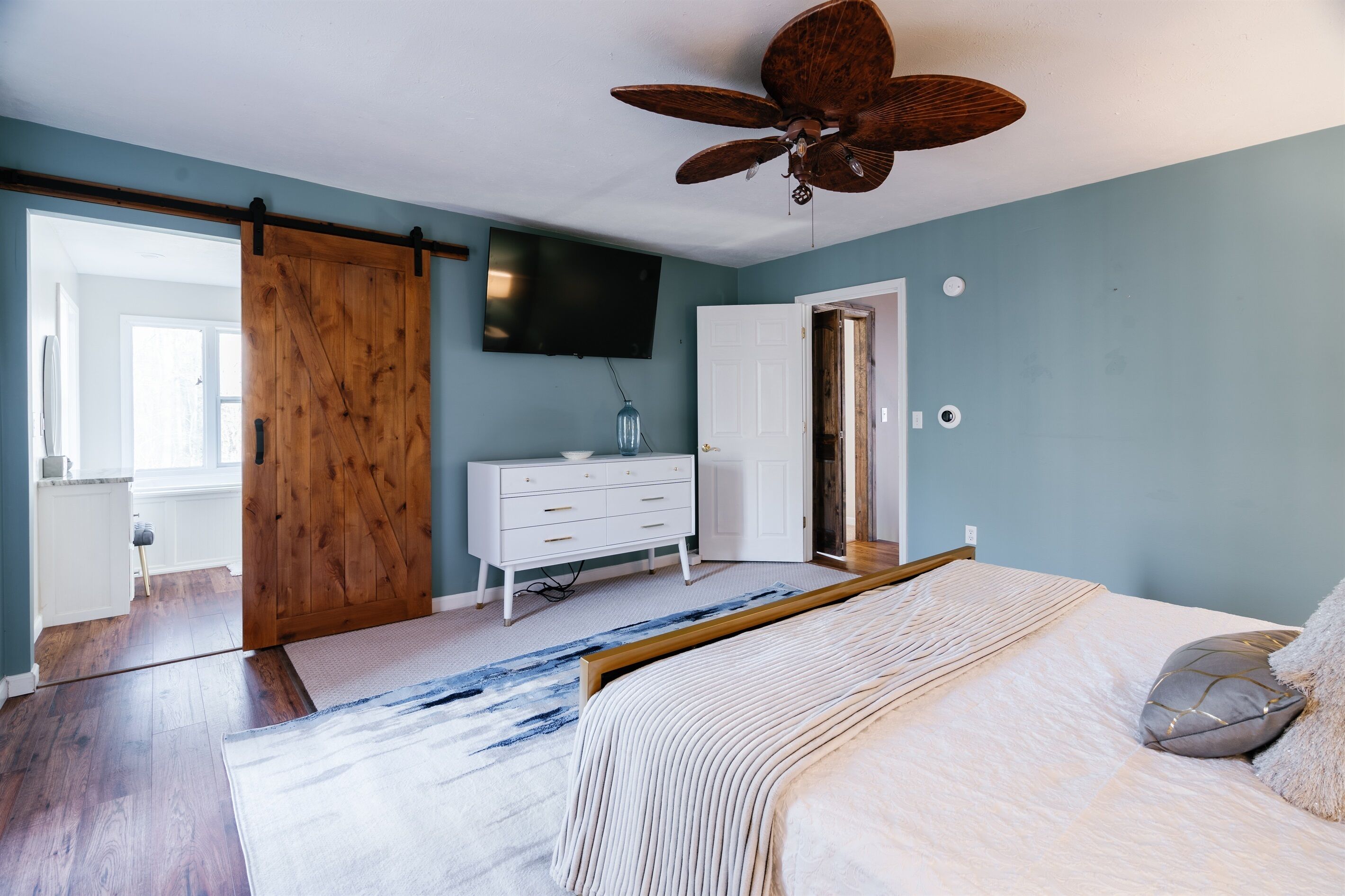A bedroom with a bed, a wooden wardrobe, a white dresser, a ceiling fan, and a TV on the wall.