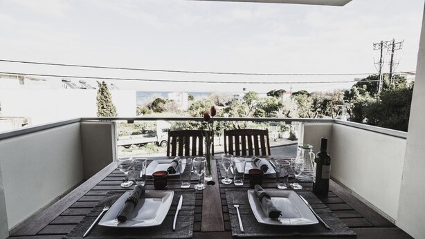 A balcony with a table set for two, overlooking a residential area with trees and buildings. - Almyra (Chios)