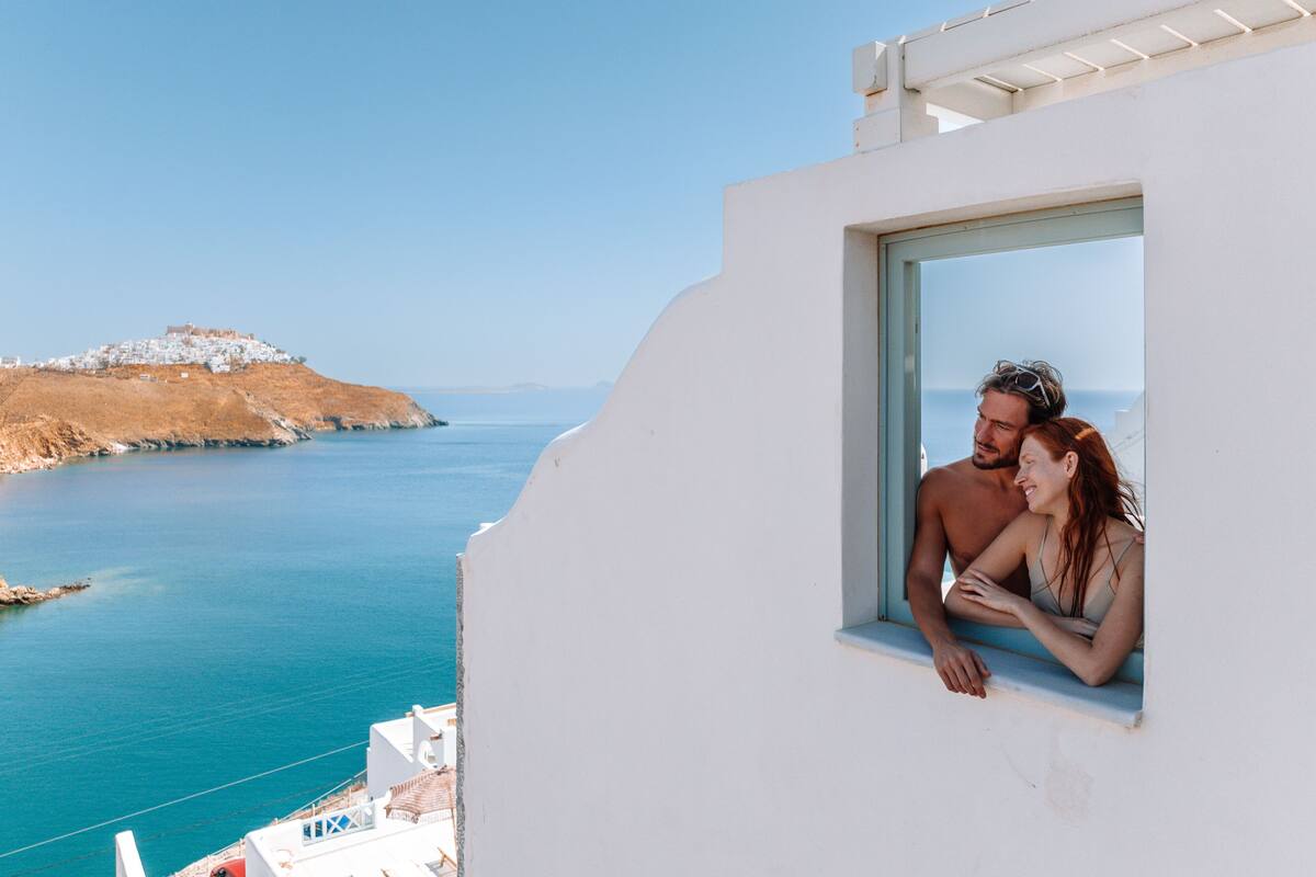 A couple on a balcony overlooking a coastal view with a white building and a distant hill.