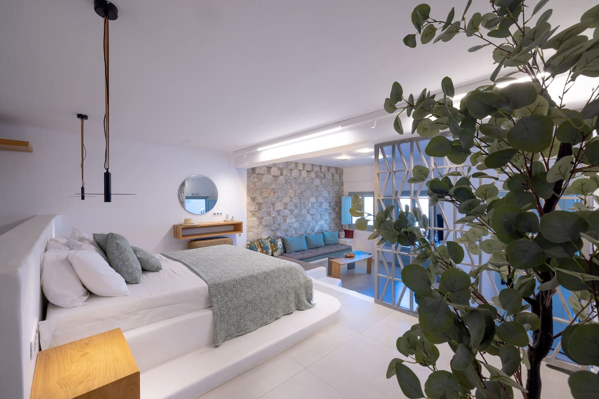 A modern bedroom with a large bed, a wooden bedside table, a stone wall, and a view of the living area.