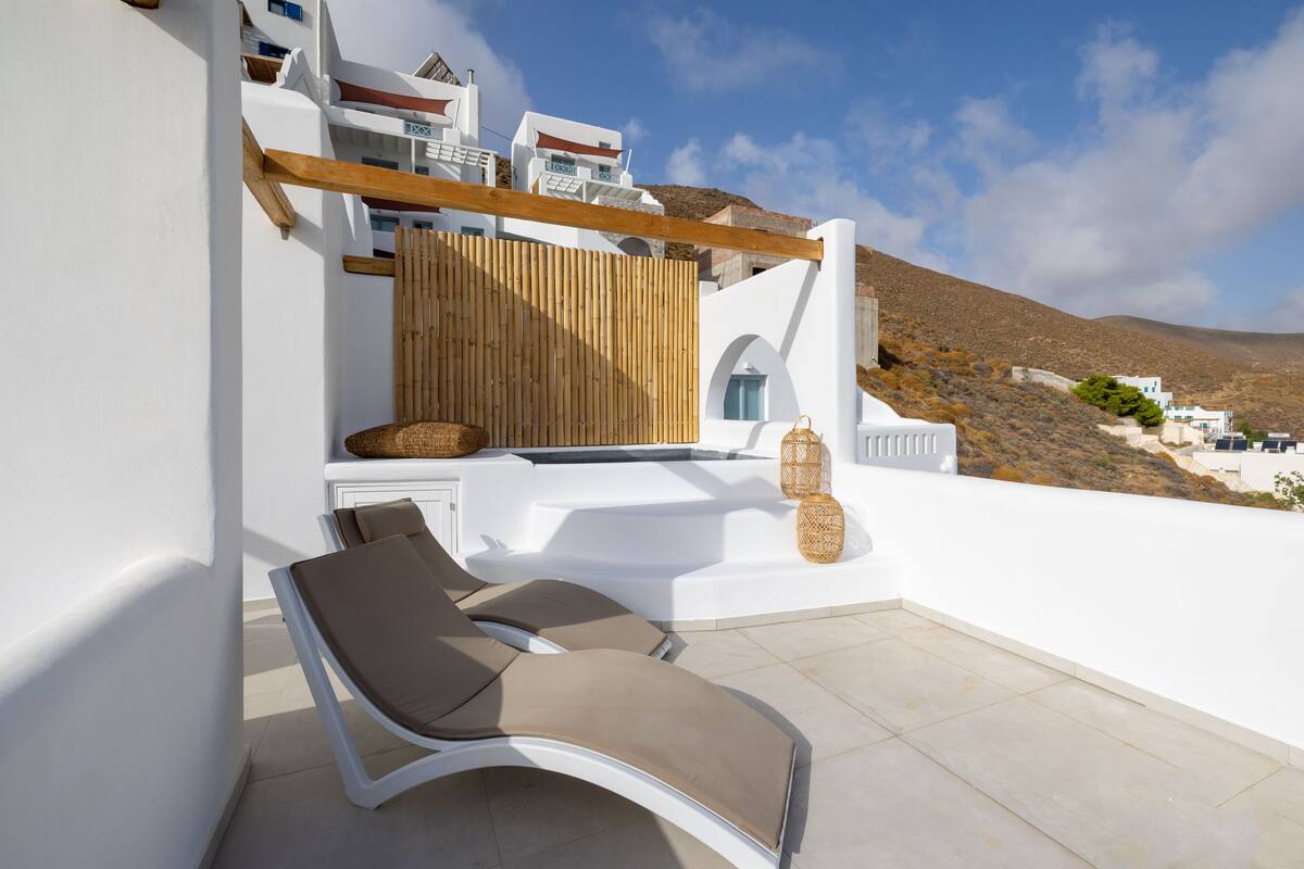 A sunlit outdoor seating area with white walls, a wooden pergola, and a view of a hillside.