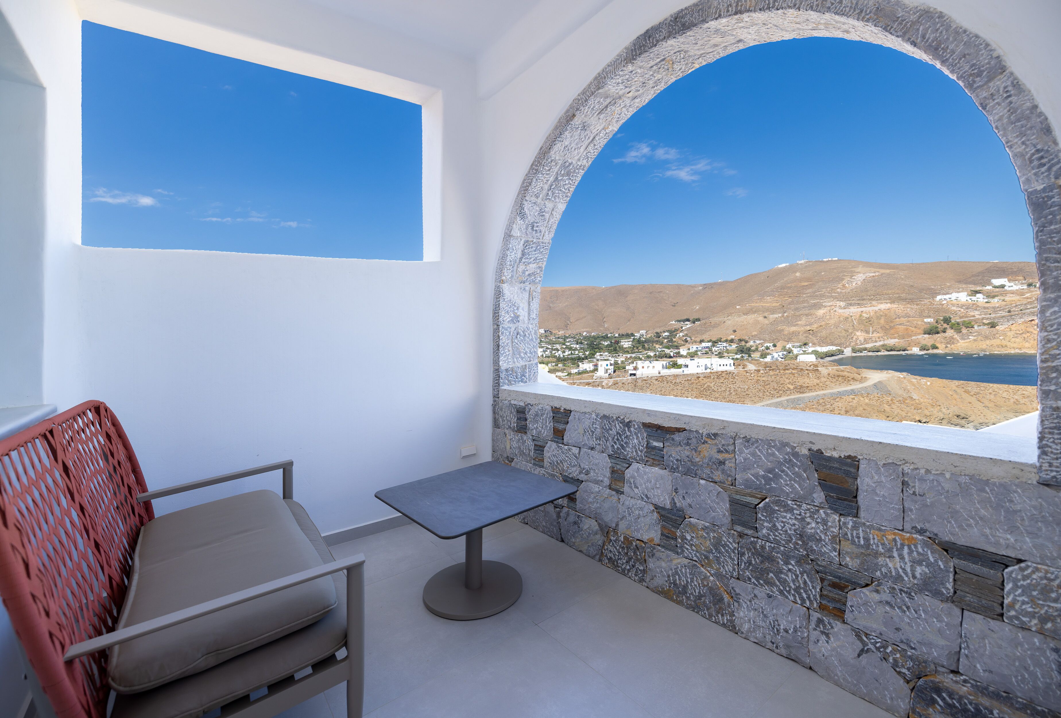 Un balcón con pared de piedra, vistas a un pueblo costero, una silla y una mesita.