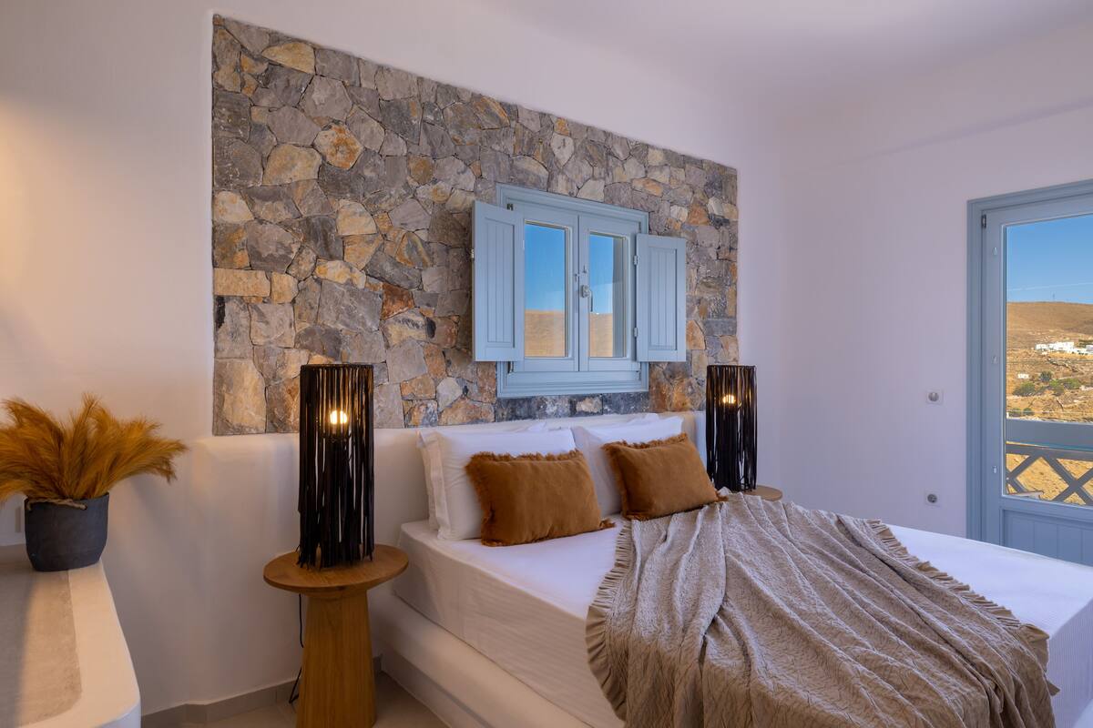 A bedroom with a stone wall, a bed with white bedding, two brown pillows, a beige blanket, and a window with a view of a landscape.