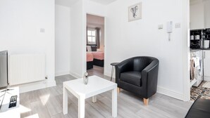 A modern living room with a white table, a black armchair, and a view into a bedroom. - Cosy and Functional 1 Bedroom Flat in London (London)