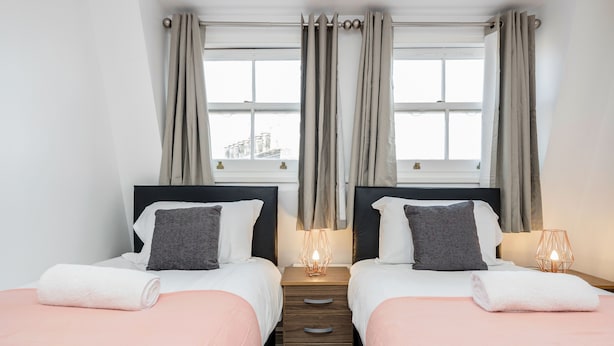 A double room with two beds, a wooden nightstand, a window with curtains, and a lamp on each side. - Cosy and Functional 1 Bedroom Flat in London (London)