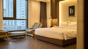 Quarto de hotel moderno com uma cama grande, uma mesa de centro redonda, uma poltrona confortável e vista para edifícios altos através de grandes janelas.