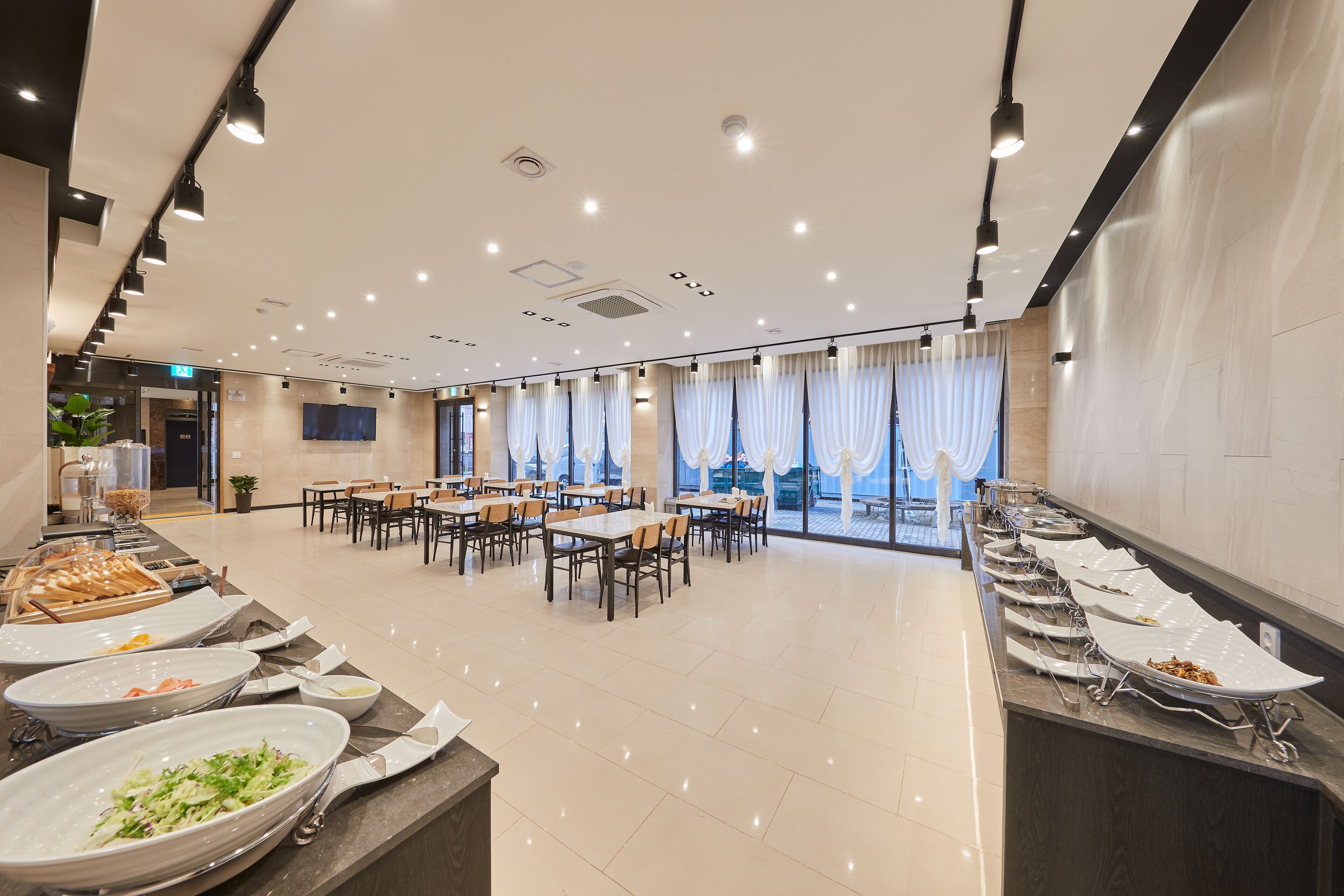 A spacious dining area with multiple tables and chairs, a buffet line with various dishes, and large windows with curtains.