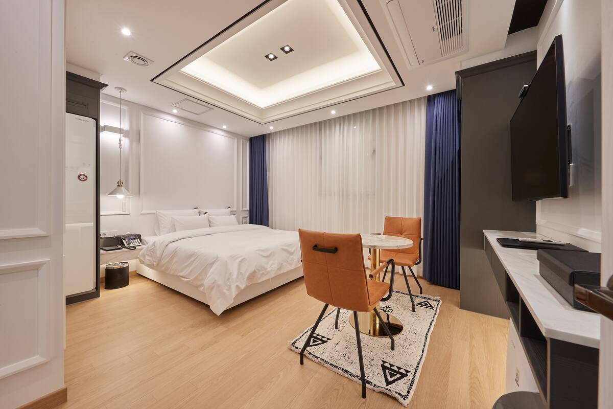 A modern hotel room with a neatly made bed, a dining area with a table and chairs, a flat-screen TV, and a ceiling light fixture.