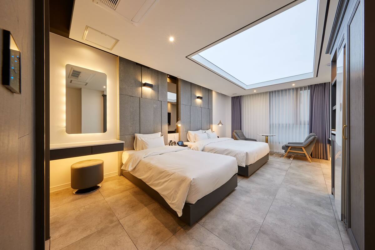 A modern hotel room with two beds, a skylight, a large mirror, and a seating area.