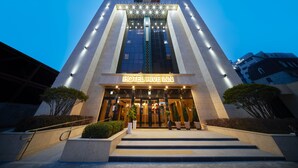 Uma entrada de hotel moderna com uma placa visível, uma escadaria que leva às portas e ambiente bem iluminado.