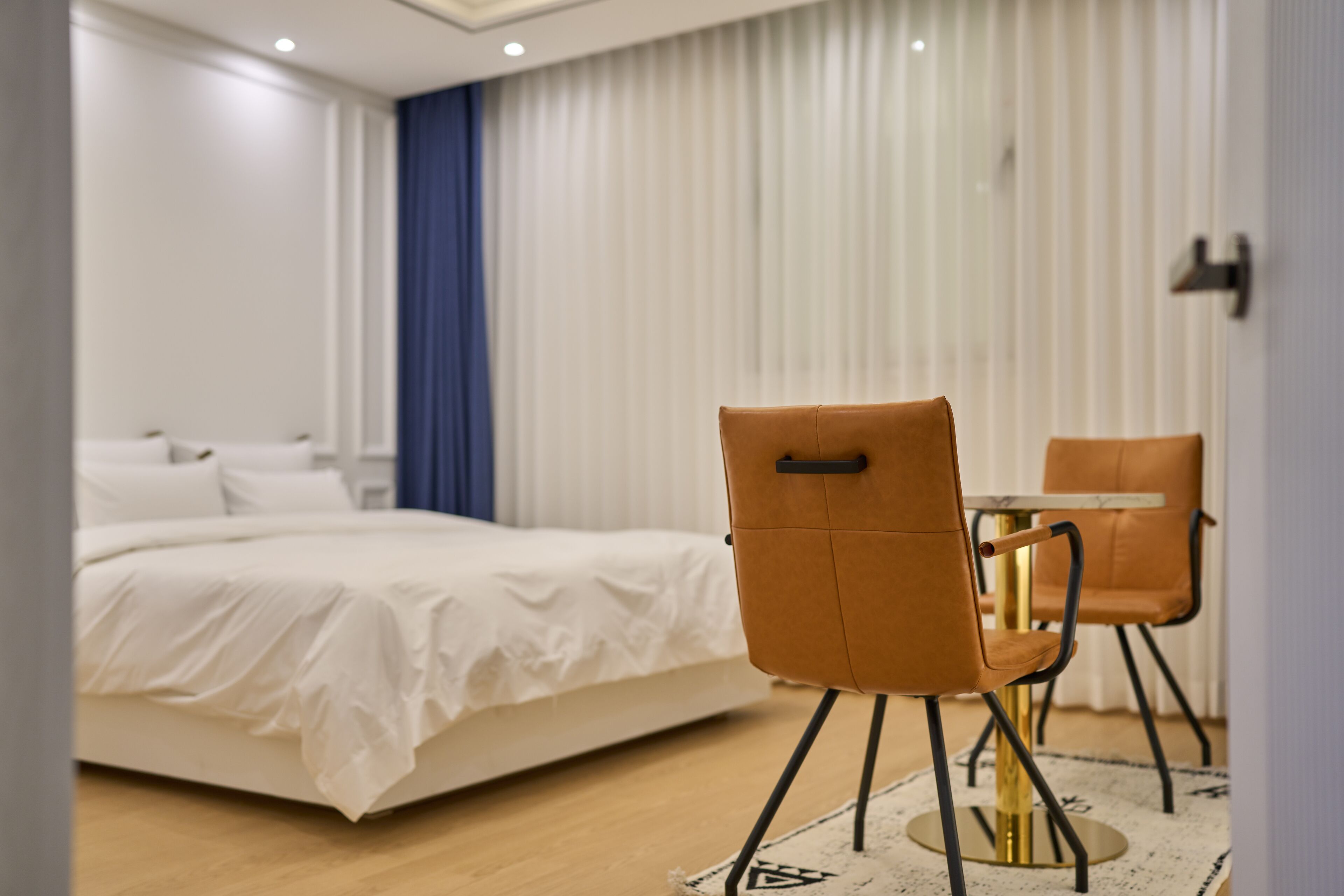 A modern hotel room with a neatly made bed, two brown chairs around a small table, and a large window with sheer curtains.