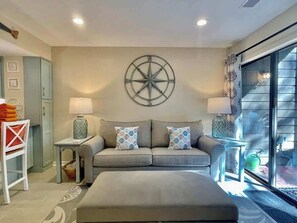 A modern living room with a sofa, two side tables, a lamp, a decorative wall piece, and a view through a glass door.