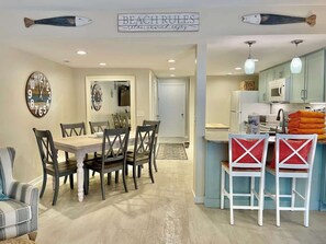 A dining area with a wooden table and chairs, a kitchen with light blue cabinets, and a sign above the dining area that reads 'BEACH RULES relax, unwind, enjoy'.