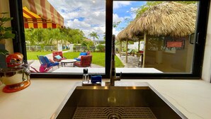 A kitchen with a stainless steel sink, a window overlooking a patio with outdoor furniture and a thatched-roof structure.