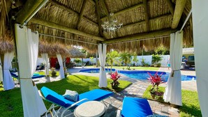 A covered outdoor seating area with blue lounge chairs and a small table, a pool, and a thatched roof structure.