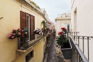 A narrow street with buildings on either side, featuring balconies with flower boxes and metal railings. - Suite 1 (Siracusa)