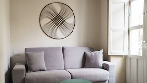 Een sofa in neutrale tinten met twee kussens, een klein rond tafeltje en een abstract kunstwerk aan de muur.