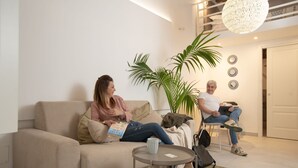 A living room with a beige sofa, a small round table, and a potted plant. - Suite 2 Standard (Syracuse)
