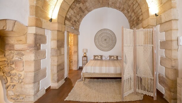 A bedroom with a stone archway, a bed with a patterned headboard, a round wall hanging, and a macramé curtain. - Suite 4 Loft (Siracusa)