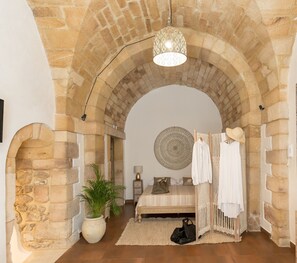 A bedroom with stone arches, a bed with a patterned bedspread, a hanging light fixture, and a large decorative plate on the wall. - Suite 4 Loft (Siracusa)