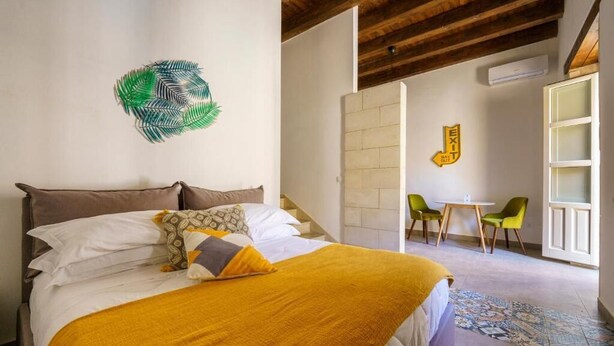 A bedroom with a bed, a green wall art piece, a yellow exit sign, and a small table with chairs. - Suite 5 Superior (Siracusa)