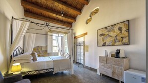 A bedroom with a four-poster bed, a bedside table with a lamp, a dresser, and a refrigerator. - Suite 7 Deluxe (Siracusa)