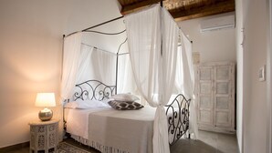 A bedroom with a four-poster bed, a bedside table with a lamp, a patterned rug, and a wooden ceiling beam. - Suite 7 Deluxe (Siracusa)