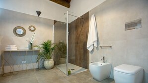 A modern bathroom with a glass shower enclosure, a freestanding bathtub, and a toilet. There are decorative mirrors and potted plants on a vanity. - Suite 7 Deluxe (Siracusa)