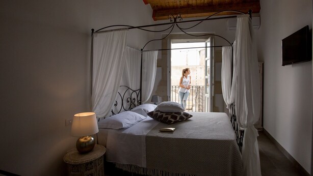 A bedroom with a four-poster bed, a bedside lamp, and a view of a balcony. - Suite 7 Deluxe (Siracusa)
