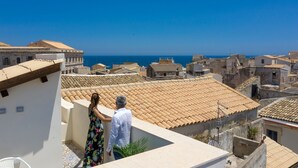 Una terraza en la azotea con vistas al mar, con tejados de tejas rojas y un cielo azul despejado.