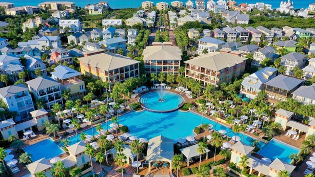 Vista aérea de un resort con varias construcciones, una piscina central con fuente y palmeras alrededor.