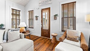 Una habitación acogedora con suelo de madera, dos sofás blancos, una puerta de madera y ventanas grandes con persianas.