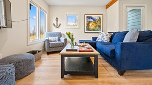 Un salon avec un canapé bleu, un fauteuil à motifs, une table basse avec des livres et un vase, et de grandes fenêtres.